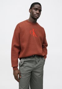 Rūsas krāsas džemperis ar apaļu izgriezumu un sarkanu izšūtu "CK" logotipu. Kombinēts ar gaiši pelēkām, brīvā fitā biksēm ar aukliņu.