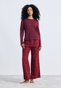 Kvinde med krøllet hår står barfodet iført en langærmet maroon top og røde ternede brede bukser foran en ensfarvet baggrund.