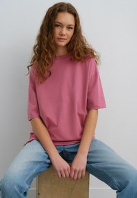Lyserød oversized t-shirt med korte ærmer, parret med lyseblå jeans. Stoffet ser blødt ud med en glat tekstur. Sittende på en træboks.