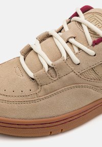 Tenis din suede bej cu șireturi albe, accente burgund și detalii perforate pe vârf. Formă rotundă și talpă din cauciuc cu textură.