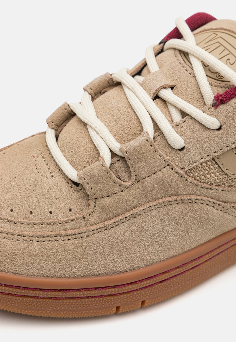 Sneaker in suede beige con lacci bianchi, dettagli in bordeaux e traforature sulla punta. Forma arrotondata e suola in gomma con texture.