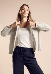 Harmaa napillinen paita valkoisen toppatakin päällä, yhdistettynä tummansinisiin joustaviin housuihin. Silkkinen kangas, yksinkertainen muotoilu.