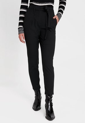 Vrouw draagt zwarte, hooggetailleerde tapered broek met strikceintuur, gestreept top met lange mouwen en glimmende zwarte enkellaarzen, staat met één hand in haar zak.
