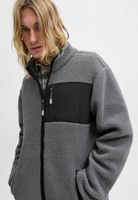 Grijze fleece jas met een zwart paneel op de borst, ritszakken en een textuurstof. Het ontwerp heeft een staande kraag en een relaxed fit.