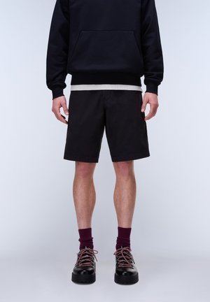 Mann trägt schwarze Shorts, schwarzen Sweatshirt über grauem Hemd, kastanienbraune Socken und schwarze Schuhe mit pinken Schnürsenkeln vor weißem Hintergrund.