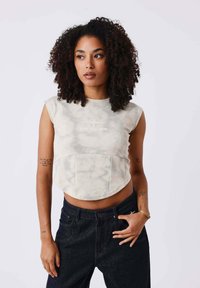 Top court sans manches dans un motif tie-dye beige clair avec un design arrondi et un panneau avant, associé à un jean foncé taille haute.