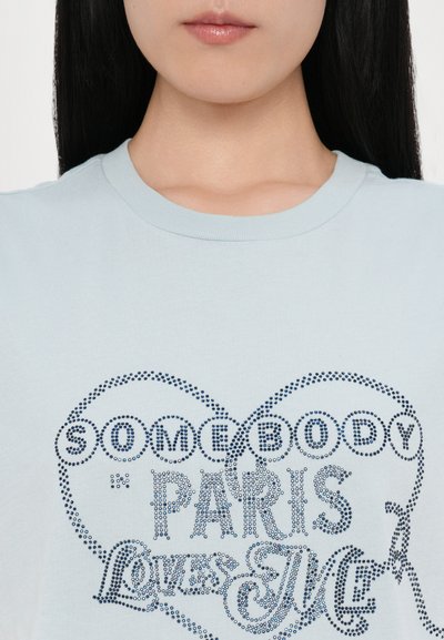 Svijetloplava pamučna majica s dizajnom srca i tamnoplavim zakovanim tekstom "SOMEBODY PARIS Loves ME." Klasični okrugli izrez.