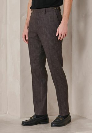 Homme portant un pantalon slim à carreaux marron avec des mocassins noirs, debout contre un mur beige.