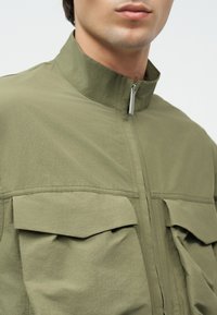 Κοντινό πλάνο ενός ατόμου που φοράει ένα χακί μπουφάν με όρθιο γιακά, κλείσιμο με φερμουάρ και δύο μεγάλες εμπρόσθιες τσέπες με καπάκι.