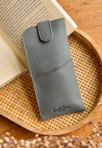 Šedý kožený obal na telefon s elegantním designem, který má přední kapsu a prošívané detaily, ležící na tkaném ubrusu vedle knihy.