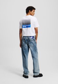 Witte katoenen t-shirt met horizontale zwarte en blauwe strepen op de rug, gecombineerd met lichtblauwe denim jeans en zwarte schoenen.