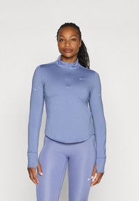 Langermet, lyseblå treningspullover med halv-glidelås i kragen. Har et Nike-logo på brystet og tommelhull i mansjettene.