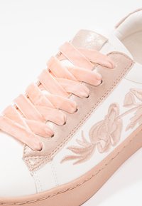 Baskets blanches avec lacets en velours rose, œillet métallique rose et motif floral brodé sur le côté.
