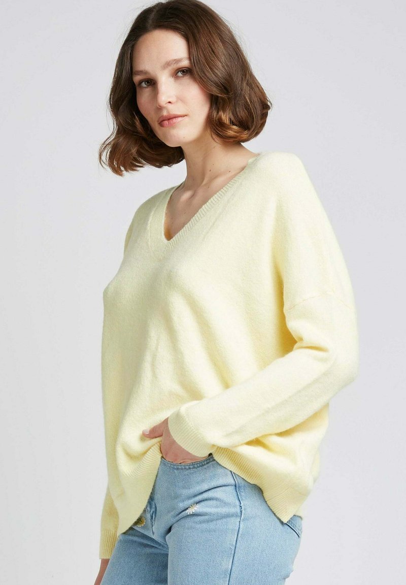 Pull à col en V, à manches longues, en matériau jaune doux avec une coupe décontractée et des poignets côtelés. Associé à un jean bleu clair avec des accents floraux.