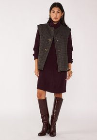 Abito burgundy a costine abbinato a un gilet senza maniche a quadri. Stivali in pelle marrone lunghi con un leggero tacco. Accenti in hardware color oro.