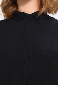 Chemise noire à boutons avec col, boutons sur le devant et poche poitrine ; texture lisse avec un design classique.