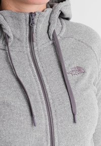 Šedá fleecová mikina s kapucí, horizontální pruhy, přední zip, dvojité stahovací šňůrky a malé logo na hrudi. Měkká textura, moderní střih.