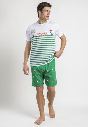 Hombre con camiseta de rayas blancas y verdes del equipo de golf Snoopy y pantalones cortos verdes con patrón de Snoopy, de pie descalzo sobre un fondo blanco.