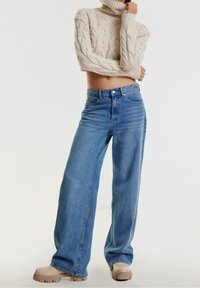 Suéter de punto trenzado color beige claro con cuello alto; jeans de mezclilla azul de tiro alto y pierna ancha; zapatos beige con suela gruesa.