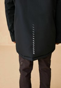 Schwarze wasserdichte Jacke mit langem Schnitt, ausgestattet mit einem reflektierenden vertikalen Streifen und kontrastierendem dunklem Stoff. Die Hose ist braun und hat Taschen.