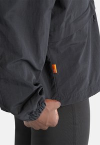 Giacca nera leggera con finitura strutturata, polsini elasticizzati e dettaglio con etichetta arancione vicino all'orlo. Mano posata sulla vita.