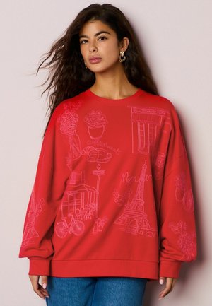 Bluză roșie cu mâneci lungi, având motive pariziene brodate roz, inclusiv Turnul Eiffel, croissante și ghivece cu flori.