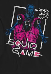 Black t-shirt featuring a graphic design with a blue figure and pink hooded characters. Text reads "BEGIN THE NEXT GAME" and "SQUID GAME."