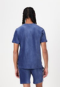 Kék cordura póló, rövid ujjakkal, függőleges csíkokkal és kontrasztos oldalsó részletekkel. Sima textúra, laza fazon.