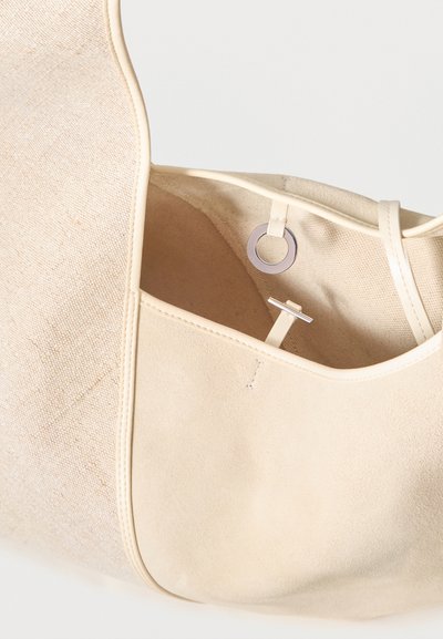 Gros plan sur un cabas en toile couleur crème ouvert, avec une poche bordée de cuir et une fermeture métallique à anneau et barre.