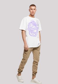 White oversized t-shirt with purple dragon graphic, paired with beige cargo pants and white sneakers. Casual outfit with relaxed fit.