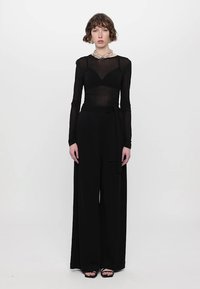 Black jumpsuit with long sleeves and a sheer bodice, featuring a tied waist, wide legs, and a textured finish. Complements with chunky jewelry.