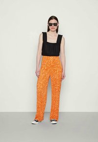 Mulher vestindo uma blusa preta sem mangas, calças largas com padrão laranja brilhante, óculos de sol pretos e ténis preto e branco, em pé contra uma parede simples.