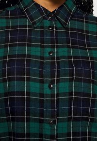 Chemise à carreaux verts et noirs avec col, devant boutonné et surpiqûres blanches. Fabriquée en tissu texturé.