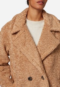 Beige dubbelgeknoopte jas gemaakt van textuurrijke stof, met een brede kraag en donkere knopen. Daaronder is een crèmekleurige trui-halslijn zichtbaar.