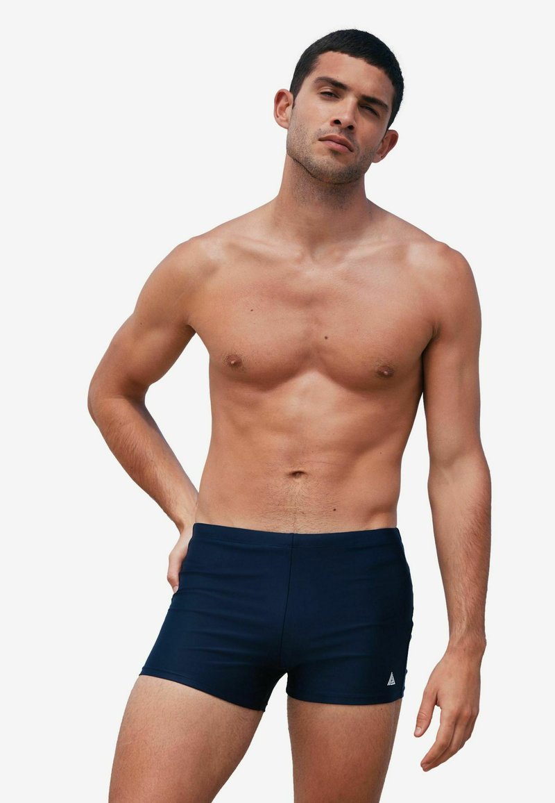 Uomo senza maglietta con capelli corti e scuri che indossa pantaloncini da bagno blu navy aderenti, in piedi davanti a uno sfondo bianco uniforme.