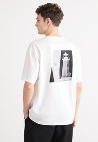 Weißes Baumwoll-T-Shirt mit einem grafischen Druck eines Leuchtturms in Schwarz und Grau auf der Rückseite. Kurze Ärmel und eine lockere Passform.