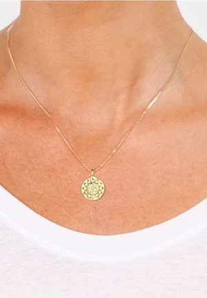 Collier en or avec pendentif circulaire présentant un symbole Om, porté autour du cou d'une personne portant une chemise blanche.