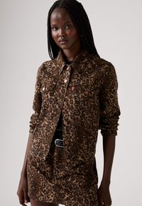 Veste en denim à imprimé léopard avec un col, fermeture à boutons sur le devant et poches poitrine. Présente une coupe structurée et un motif de couleurs marron et noir.