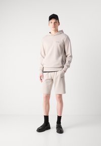 Haut à capuche beige clair et shorts assortis, fabriqués dans un matériau doux avec un logo subtil. Porté avec des chaussettes noires et des chaussures noires imposantes.