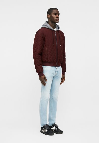 Homme debout portant une veste matelassée bordeaux avec une capuche grise, un jean bleu clair et des baskets noires à rayures blanches, sur un fond uni.