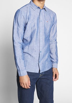 Man draagt een lichtblauw overhemd met lange mouwen en borstzak, en donkere blauwe jeans, staat met ontspannen armen tegen een witte achtergrond.