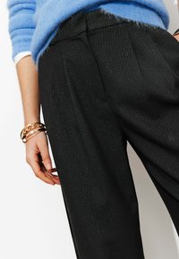 Pantalon noir plissé à fines rayures discrètes, associé à un pull bleu clair. La tenue comprend plusieurs bracelets dorés au poignet.