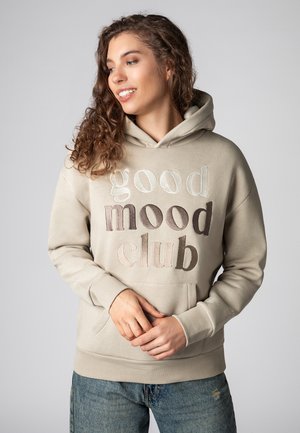 Beige hoodie met de tekst "good mood club" in verschillende borduurskleuren. Voorzien van een kangoeroezak en geribde boorden. Gemaakt van zacht katoen.