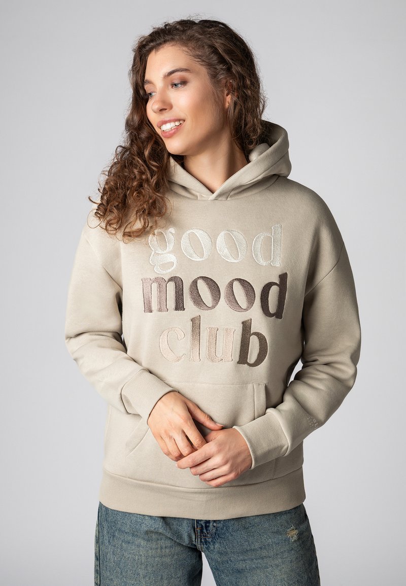 Beige hoodie met de tekst "good mood club" in verschillende borduurskleuren. Voorzien van een kangoeroezak en geribde boorden. Gemaakt van zacht katoen.