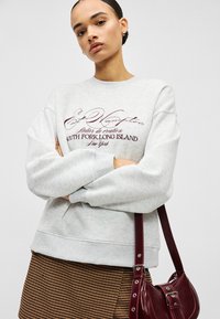 Pelēka kokvilnas džemperis ar izšūtu bordo tekstu. Aprīkots ar apaļu kaklu un ribām ap atlokiem. Kombinēts ar brūnu pundurkoku raksta svārkiem un bordo somu.