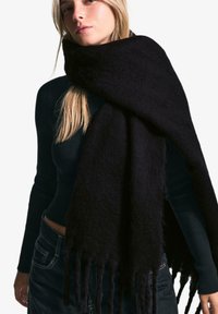 Écharpe noire en tissu doux et texturé, avec des bords frangés. Portée drapée autour du cou sur un haut ajusté à côtes.
