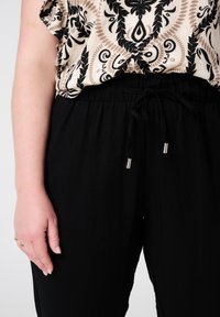 Zwarte broek met elastische tailleband en striksluiting, met een zachte textuur. De blouse is lichtbeige met een zwart paisleypatroon.