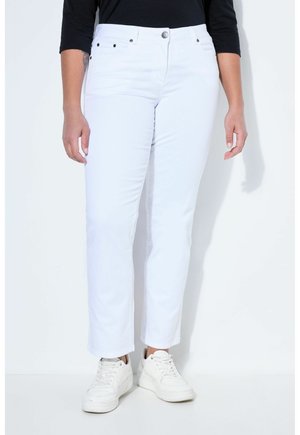 Donna che indossa jeans bianchi a gamba dritta, maglia nera a maniche lunghe e sneakers bianche, in piedi contro uno sfondo bianco uniforme.