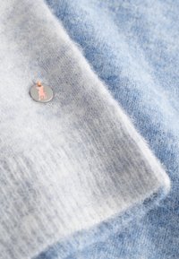 Zachte, fluweelachtige breisels in lichtblauw en lichtgrijs met een klein rond metalen label waarop een roze hondensilhouet is bevestigd.