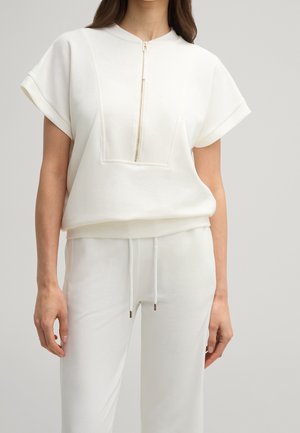 Kobieta w białej bluzce z krótkim rękawem z zamkiem z przodu i dopasowanych spodniach ze ściągaczem, stojąca na jednolitym jasnym tle.